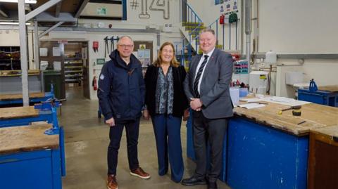 Left to right there are Chris Ambler, CEO Jersey Electricity, Jo Terry-Marchant, Principal Highlands College, Rob Ward, Minister for Education and Lifelong Learning. The three of them are standing inside a workshop. 