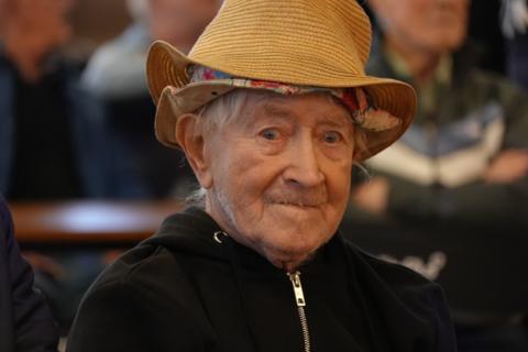 Ron "Ginger" Parsons looking at the camera, watching a performance at his 103rd birthday party. He is wearing a straw hat over the top of a floral bucket hat. He has a black zipped up hoodie on.