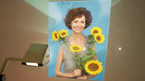 An A1 image of a woman with short curly hair, holding sunflowers, being projected onto a wall, she is smiling and looking straight at the camera 