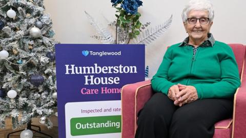 An elderly woman with short white hair and black-rimmed glasses is sitting on a red sofa chair. She has her hands together on her lap and is smiling to the camera. She is wearing a green zip cardigan and black trousers. To her right is a Christmas tree with silver decorations and a large sign that reads 'Humberston House care home inspected and rated outstanding.