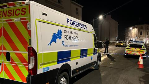 A forensics van and a police cordon on a road in Plymouth. There is an officer and cars behind the cordon.