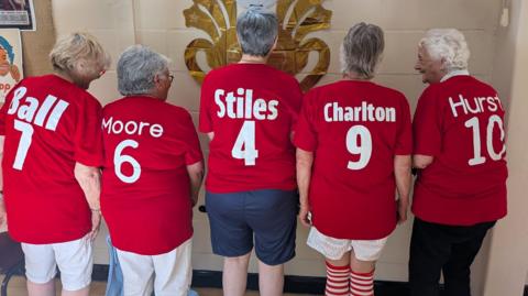 group of five women, wearing football shirts, with their backs to the camera. 