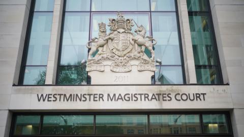 The exterior of Westminster Magistrates Court, with its name written above the entrance. Above the text, there's a large decorative coat of arms mounted on the building. The coat of arms shows a shield supported by a crowned lion on the left and a unicorn on the right, topped with a crown and a banner below that reads "DIEU ET MON DROIT."