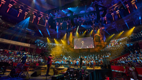 A large concert hall with multi coloured lights is filled by a huge orchestra