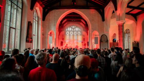 People standing in church which is being used as a music venue. There is a stage set up within a church alcove and a band is playing guitars. The stage is lit up with a red light.