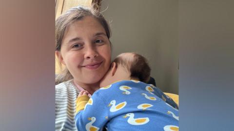 Jessica Kent-Hazledine cuddles her baby son, whose face is next to her neck. Kent-Hazledine is smiling. She is wearing a green and white stripey t-shirt. Her son is wearing a blue babygrow with white and yellow ducks on it.