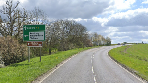The A6 between the junctions for Souldrop and Knotting Green in Bedfordshire. A green road sign is directing traffic onwards to Bedford or to the left to Knotting, Melchbourne, Yielden and to a historic church. 