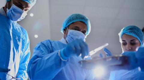 Three surgeons wearing face masks, gloves and blue scrubs look down at some surgical tools in preparation for an operation.