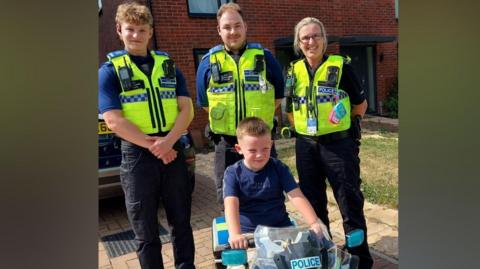 A young boy sitting on a toy police motorbike with three police officers standing behind him. They are wearing blue t-shirts and fluorescent yellow police vests