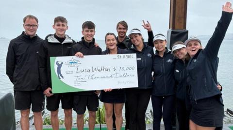 A group of people stood next to a lake. They are holding a large novelty cheque and smiling.