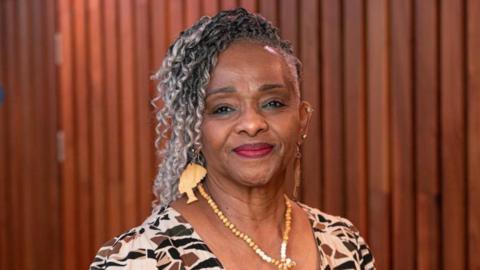 Myrle Roach is in the middle looking towards the camera with a light smile. She has long black and grey hair. She is wearing a brown and black patterned top, large wooden earrings in the shape of a woman's head, and a beaded necklace.