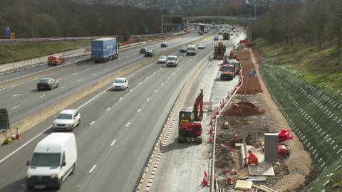 A motorway with traffic passing in both directions. On the right of the picture is a coned off area where a digger is working. There is a mound of rubble next to the digger, where a man in orange clothing works near an electrical cabinet. Next to him on the far right of the picture is an embankment covered in green netting.