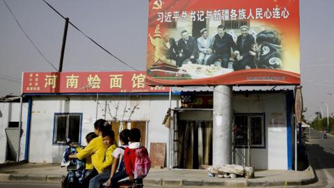 A Uyghur woman uses an electric-powered scooter to fetch school children as they ride past a picture showing China's President Xi Jinping joining hands with a group of Uighur elders at the Unity New Village in Hotan, in western China's Xinjiang region.