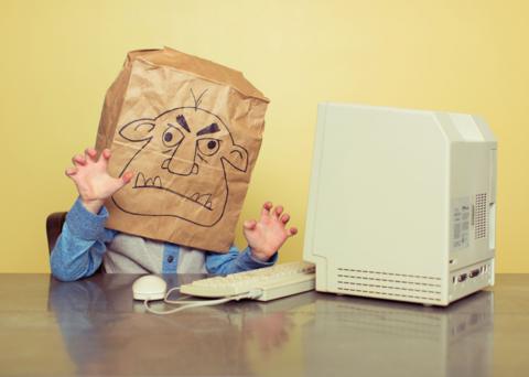 A representation of an internet troll, a child wearing a paper bag with a troll face drawn on it, sits at a desktop computor