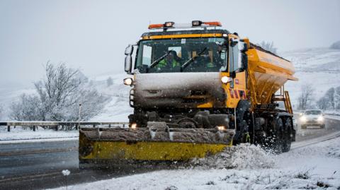 A gritter with a plough works to clear a road covered in snow