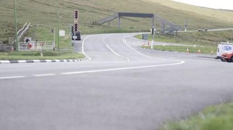 The A18 Mountain Road snaking under a pedestrian bridge over the carriageway in the direction of Douglas. The barriers and lights for the Snaefell Mountain Railway can also be seen.