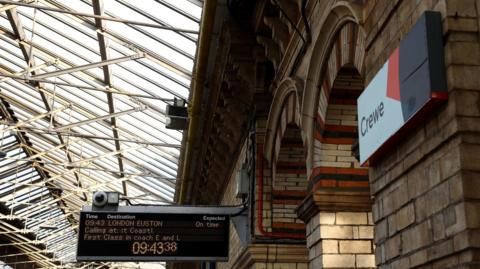 View inside Crewe railway station. There is a sign with the word Crewe on the right of the screen and a destination board for a train to London in the centre of the image.