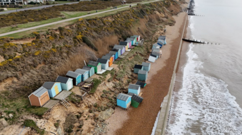 two rows of beach huts on the side of a cliff which leads down to a beach at the side of the sea. Some of the huts are leaning at unusual angles