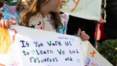 A girl is holding a banner that reads 'If you want us to learn we need resources and books'.