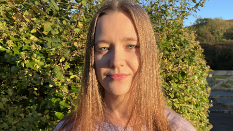 Bethan Evans, who has long straight hair, and is wearing a pink jumper, looks towards the camera, with bushes, fields and a wooden gate in the background