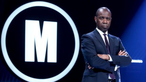 Clive Myrie standing next to the Mastermind logo