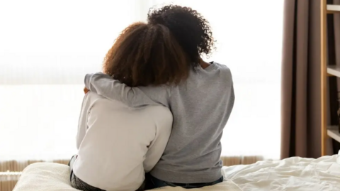 Two females sit on the edge of a bed, one with her arm around the other both facing a window. One is wearing a grey top the other a white top.