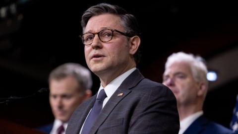 Mike Johnson looks ahead while speaking at a press conference. He has dark grey hair and wears glasses. He is wearing a dark suit, white shirt and navy tie.