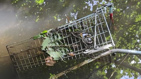 A disused, rusty trolley dumped in the Bottesford Beck. Leaves and algae are intertwined with the metal. Reflections of trees can be seen through the murky brown water.