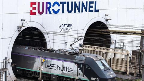 A LeShuttle train emerges from Eurotunnel - a white paneled structure with two dark tunnels. The train is dark blue and white and is attached to wires overhead.