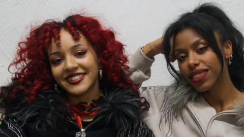Nova Twins sit on a sofa looking at the camera. Georgia South is on the left side of frame. She has red curly hair and wears black clothing, with dark red lipstick. Amy Love sits on the right side of frame. She is wearing a grey