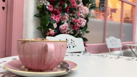 A pink cup is on a white table. It is outside a pink window. On the side of the window is a flower display. 