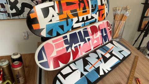 Three skate decks on a table with paint brushes and spray cans on the table - the decks are decorated in blocky graphics.