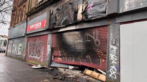 A shop front with red shutters is caved in with significant fire damage visible with black and white marks across it. 