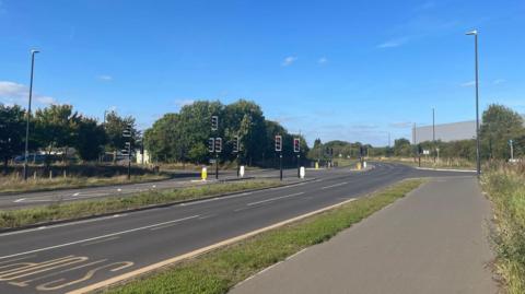 A picture of the road with traffic lights and a yellow stop sign and grass verge. Next to this is a grey pavement. 