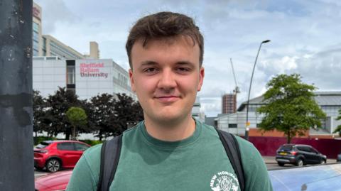 Ellis Nevitt, a young man with dark hair who is wearing a green T-shirt with backpack straps visible over his shoulders.
