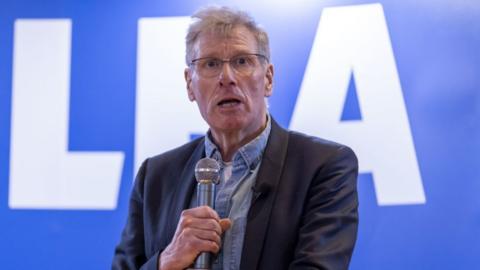 A man with grey hair speaks while holding a silver microphone in front of his mouth. He is wearing a dark suit jacket over a denim shirt. Behind him is a large blue sign with an Alba logo. 