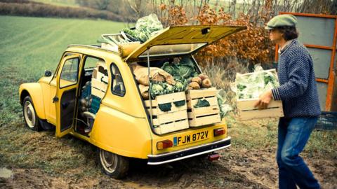 This is a photo of Guy Singh-Watson loading wooden boxes of vegetables into the boot of his yellow car. The car is parked in a green field, with a hedge in the distance.