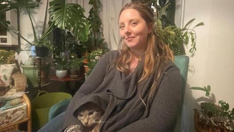 Emily Witts sits on a chair in a house with a grey cat on her lap. She is wearing a grey jumper and has long hair. There are several green plants in pots scattered on coffee tables behind her.