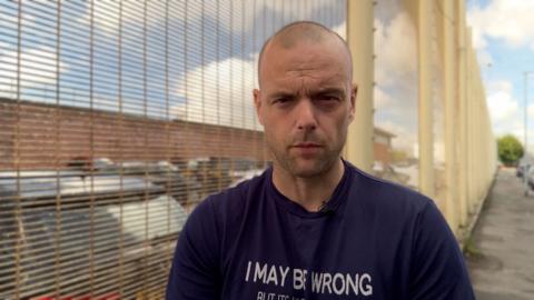 Jamie is wearing a black t-shirt and is standing next to a tall fence. He has a shaved head and stubble. 