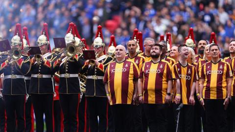 A mixed gender and aged group of people wearing Bradford City FC branded outfits, pose together in a football stadium. They stand alongside a brass band in military uniform.
