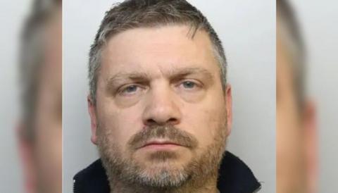 A custody image of Andrew Thorne. He has greying dark hair, and a lighter, also greying, beard and moustache. He is staring directly at the camera and appears to be wearing a dark-coloured fleece. 