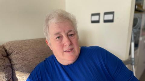 A woman with short grey hair, wearing a blue t-shirt, looking at the camera while sat on a seat in her living room. There is a crutch to the right of her.