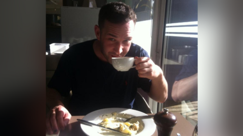 A young man with short dark hair and stubble drinks a cup of tea and eats a cooked breakfast in a cafe.