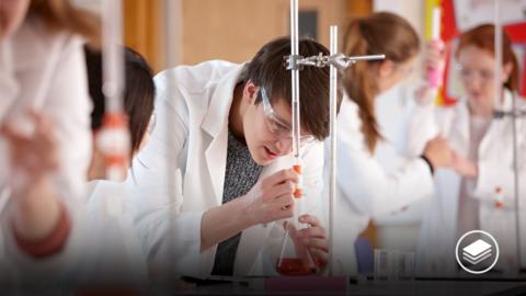 A photograph of a male student in a science lab coat, looking into a beaker with a red liquid. 