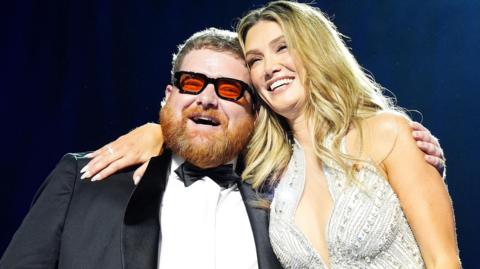 A smiling Cammy Barnes, who is wearing a dinner suit, is hugged by Delta Goodrem, who is wearing a silver sparkly dress. Cammmy has ginger hair and beard and black framed glasses with red lenses. Delta has long blonde hair.