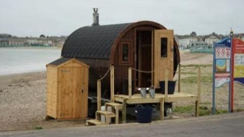 Beach and sea behind a wooden tube-shaped sauna accessed by wooden steps - the sauna door is open and a small shed stands to its left.