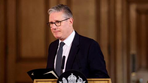 Keir Starmer standing awkwardly at a podium wearing a black suit 