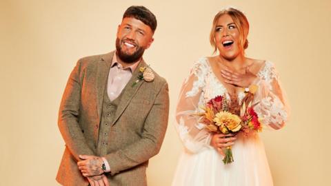 Polly and Adam in wedding outfits, with her wearing a white dress with sheer arms and a low v-neck and holding a bouquet of orange and red flowers while Adam is wearing a tweed suit in a pale brown with a pastel pink shirt and buttonhole.