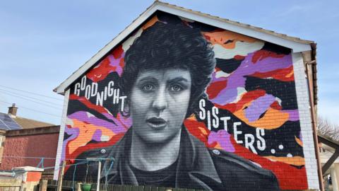 Mural of Nell McCafferty. She is in black and white with short curly hair, wearing a leather biker jacket. 'Goodnight sisters' is written either side of her. Colours of red, orange, black, purple and white are in the background.