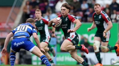 James O'Connor wearing the Leicester bottle green strip runs with the ball in hand as Finn Russell, wearing the blue, black and white of Bath prepares to tackle him. Two other Leicester players look on in the background.